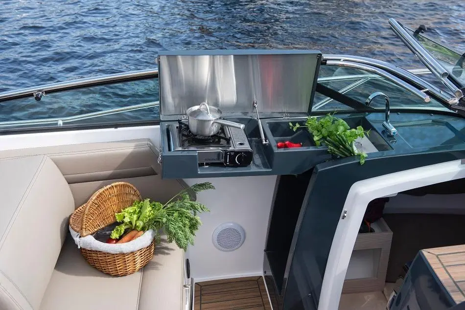 A modern boat kitchen with a gas stove, fresh herbs, and vegetables in a basket, against a backdrop of water.